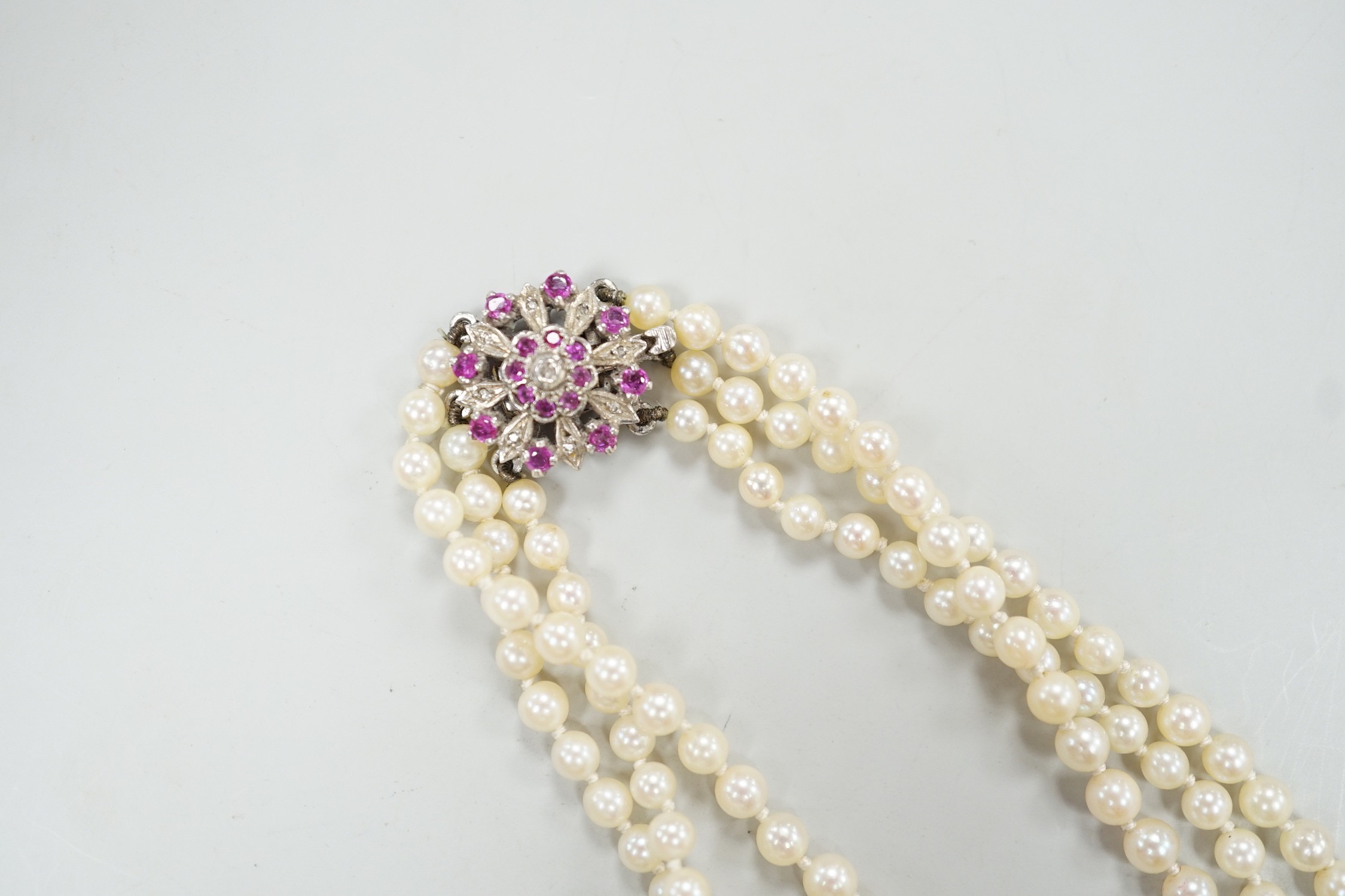 A 1960's triple strand graduated cultured pearl necklace, with 9ct white gold, ruby and diamond chip cluster set clasp, 49cm.